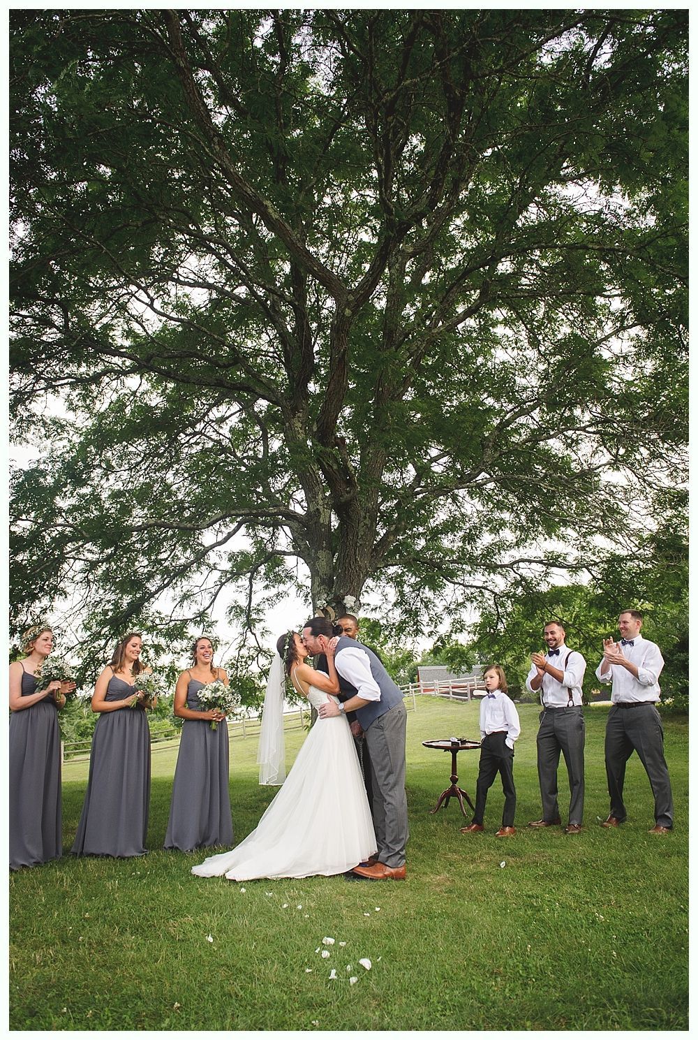 Wedding ceremony: Couple kissing under a tree, bridesmaids and groomsmen watching, outdoor setting.