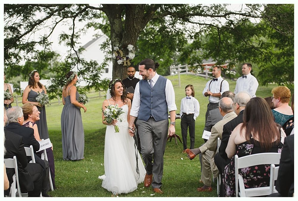 Wedding couple walking down aisle, holding hands. Guests seated outside; bridesmaids in grey dresses.