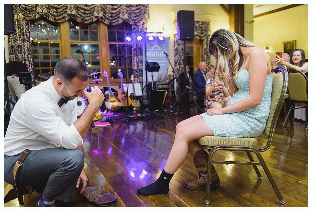 Man kneels, adjusting woman's shoe in a lit ballroom, band on stage.
