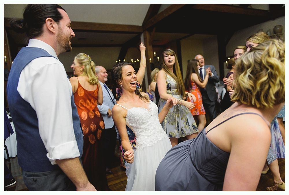 Wedding reception, bride dancing enthusiastically with guests. People are smiling, wooden ceiling.