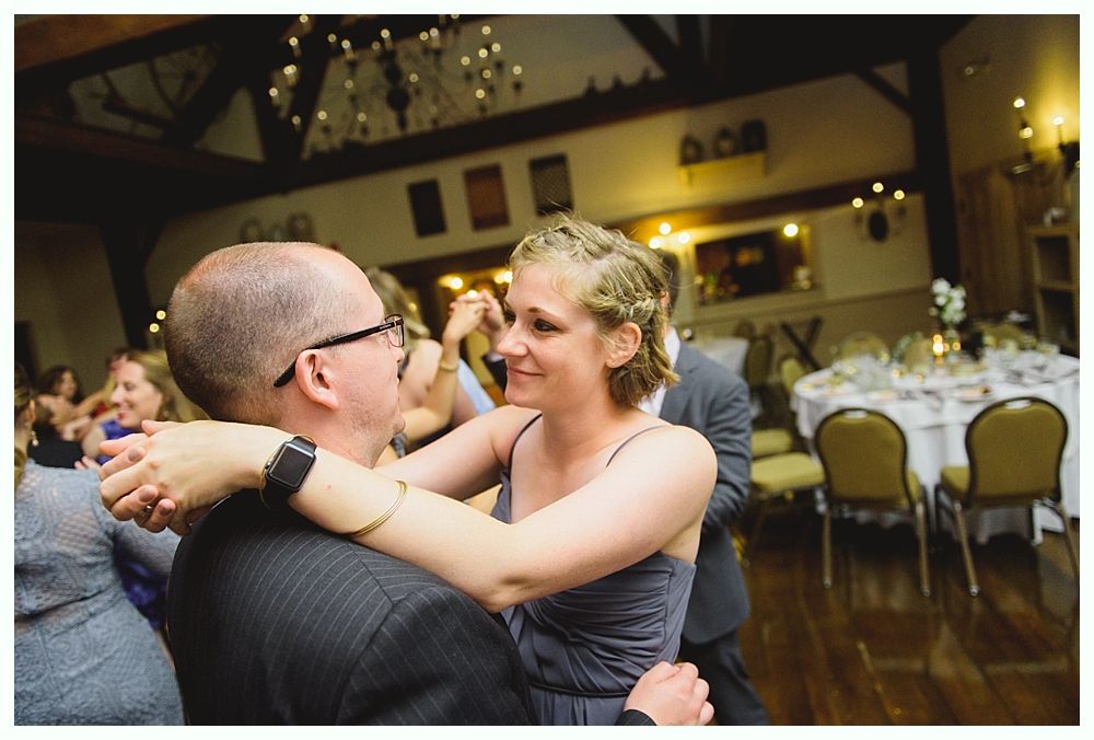 Couple dancing in a warmly lit ballroom, smiling at each other. Round tables with settings are visible in the background.