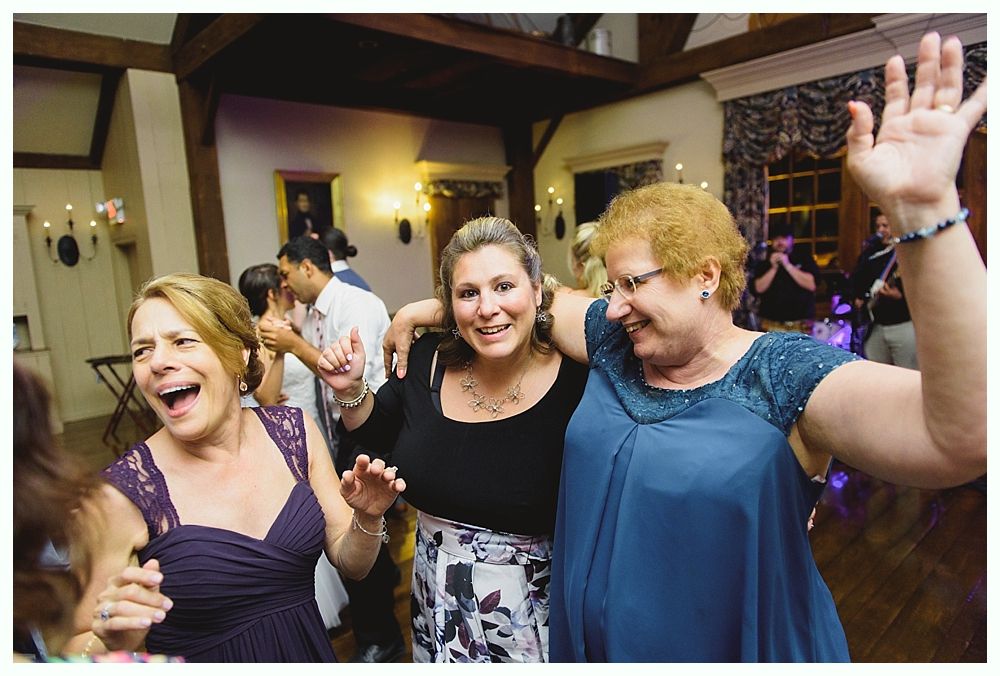 Three women dancing and laughing at a party, one with an arm raised. Dimly lit room with other people.
