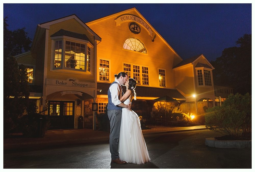 Couple kissing in front of a warmly lit yellow building at night.