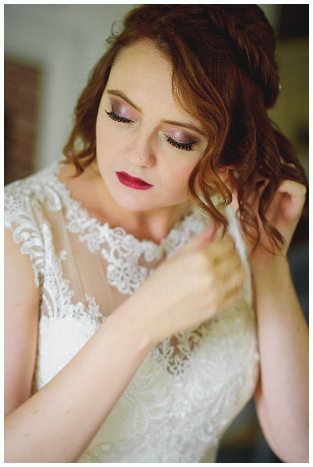 Bride adjusting her hair, wearing a white lace dress with a soft pink eye shadow, and red lipstick.