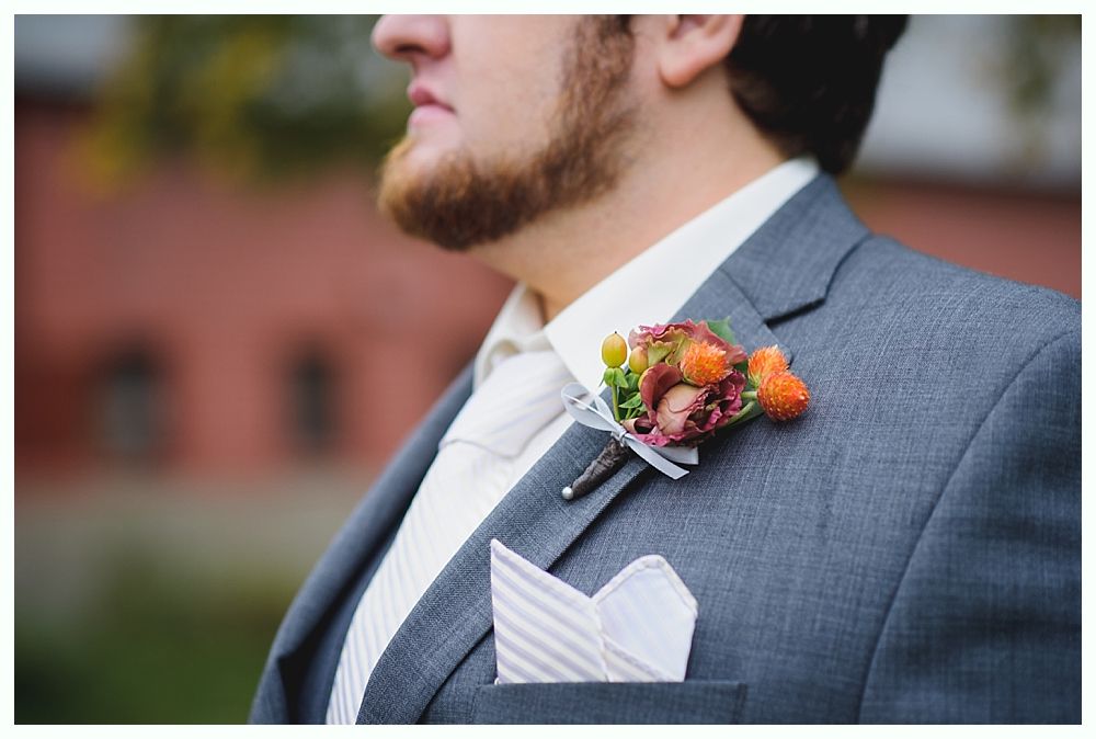 Man in gray suit with boutonniere and pocket square, outdoors, blurred red brick background.