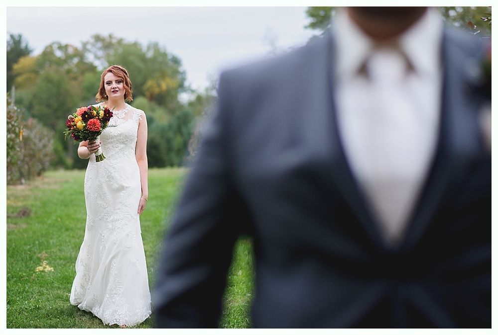 Bride in white lace dress holds bouquet, looking at blurred groom in a dark suit outdoors.