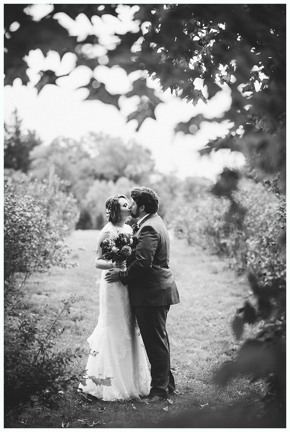 Bride and groom kissing outdoors; trees overhead, floral bouquet, black and white.