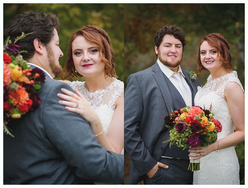 Bride and groom embrace outdoors; she wears a white lace dress and holds flowers. He wears a gray suit, smiling.