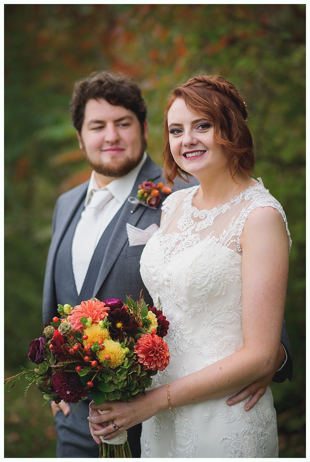 Newlyweds posing outdoors; bride in white lace dress holds bouquet. Groom in gray suit smiles.