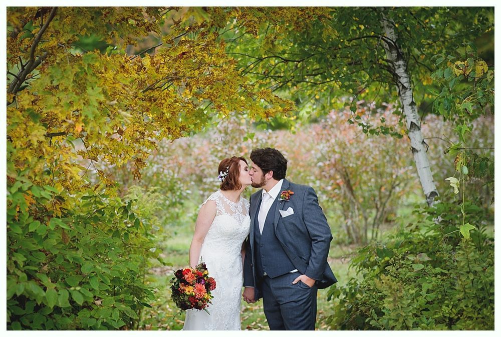 Bride and groom kiss amidst colorful fall foliage. The bride holds a bouquet.