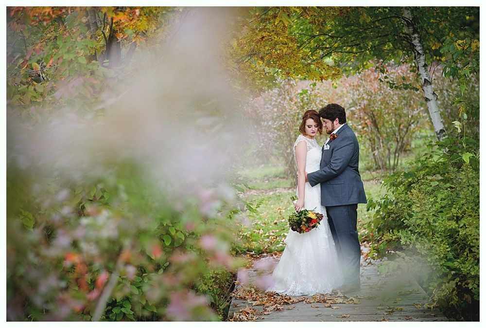 Newlyweds embracing in a garden, fall foliage, bride in white dress, groom in suit.