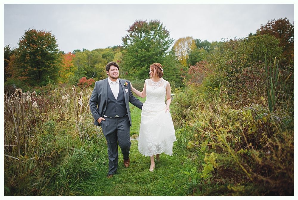 Newlyweds walk through a grassy path; groom in gray suit, bride in lace dress. Autumn foliage in the background.