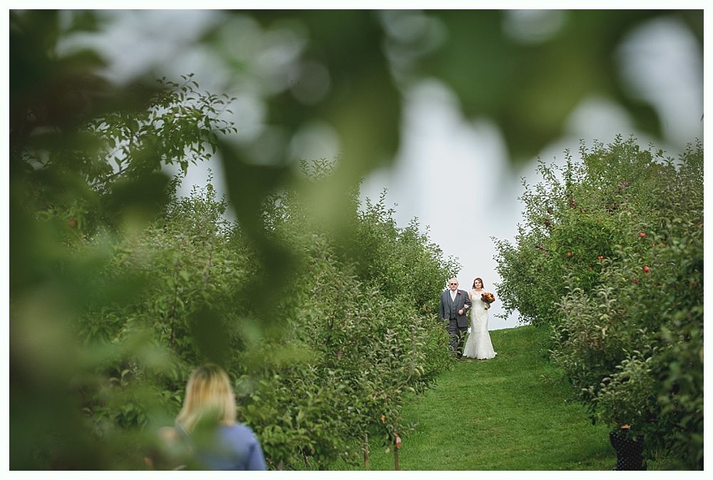 Wedding procession in a green setting. Bride walks down the aisle with her father, others watch.