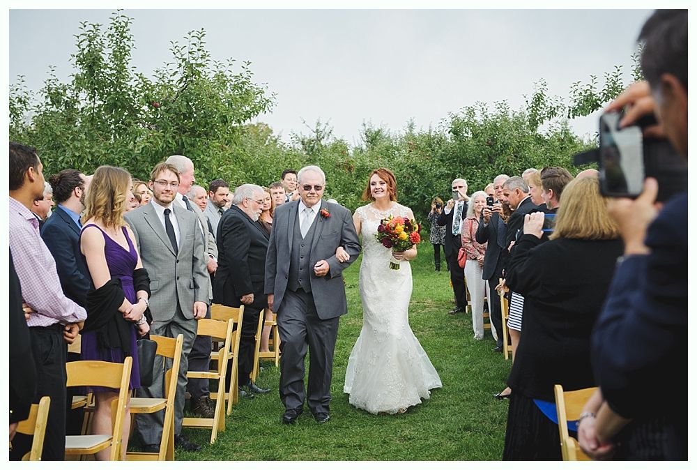 Bride in lace gown walks down aisle with her father, surrounded by guests at outdoor ceremony.