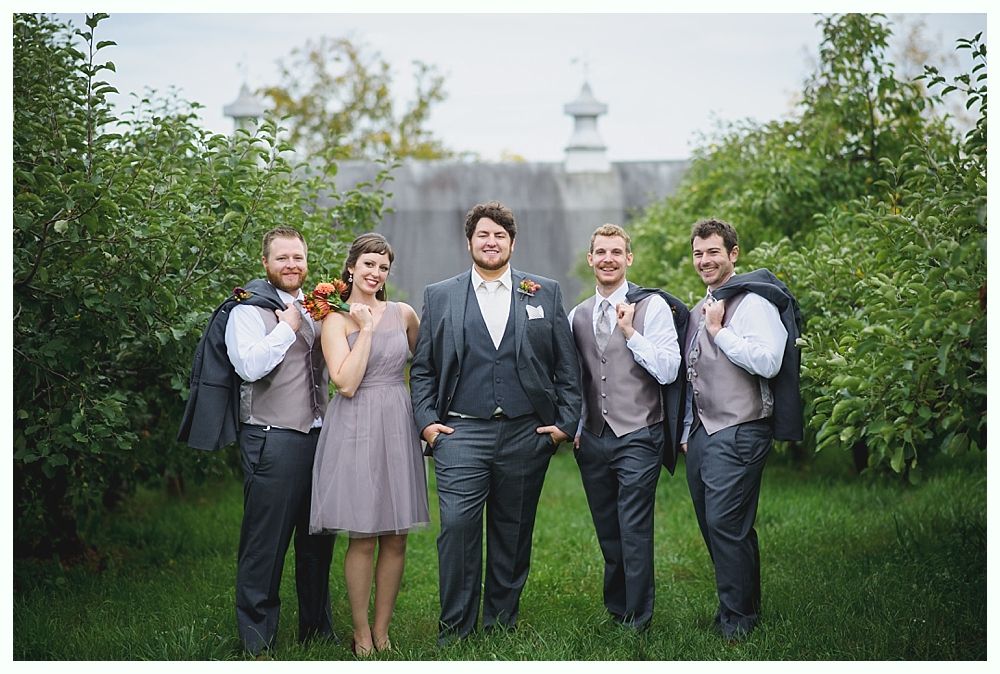 Wedding party poses outside, building in background. Bride and groomsmen in formal wear. Green trees.