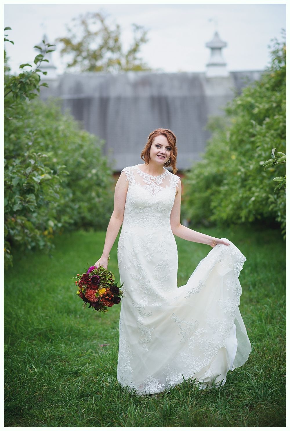 Bride in white dress, holding bouquet, poses in front of a barn.