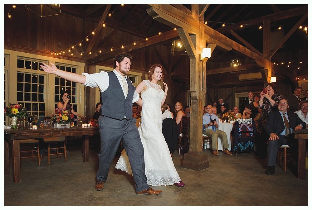 Bride and groom dancing, barn reception. Groom with arms outstretched, bride singing. Guests seated at tables.