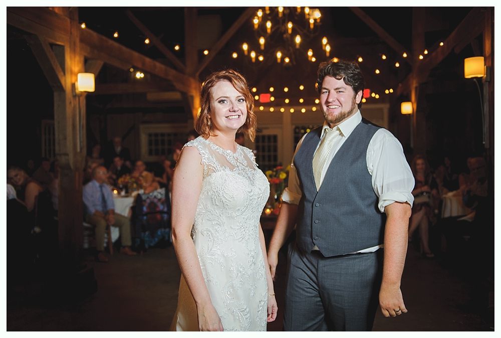 Bride and groom smiling at their wedding reception, beneath string lights and exposed wooden beams.