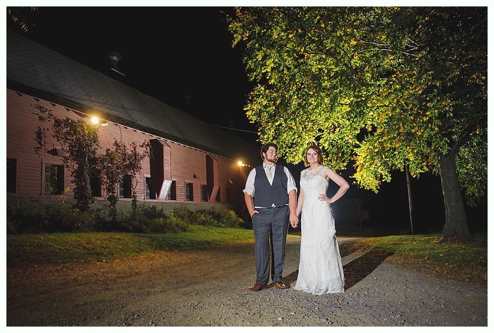 Couple holding hands on a gravel path at night, in front of a barn and tree lit by warm light.