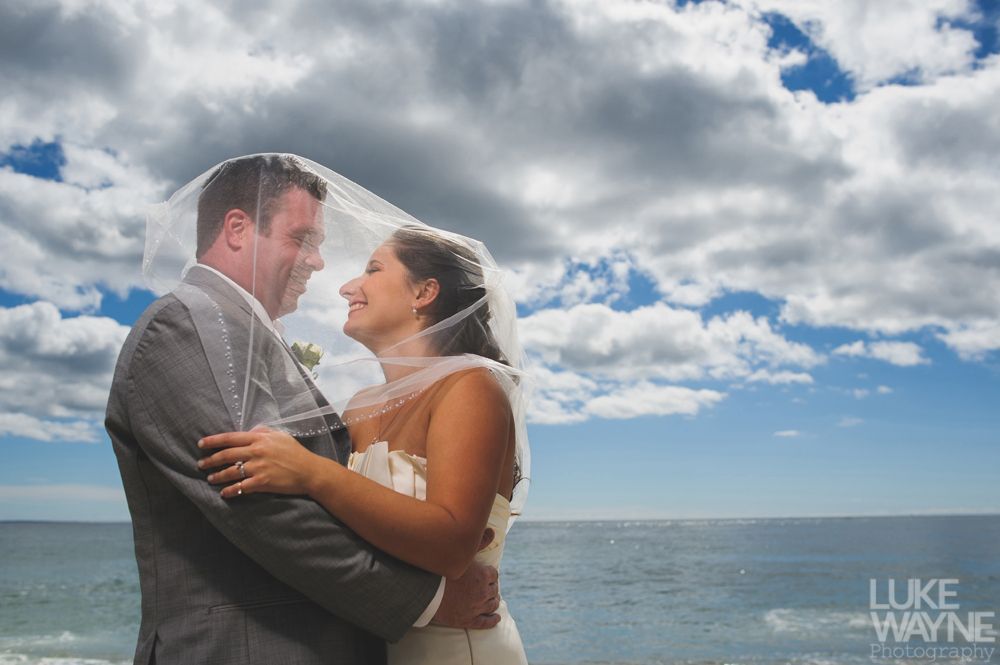 Couple embraces under a veil on beach; cloudy sky, ocean in background.