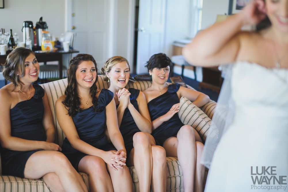 Bridesmaids watch bride adjust veil, smiling and laughing, in a room with a sofa.