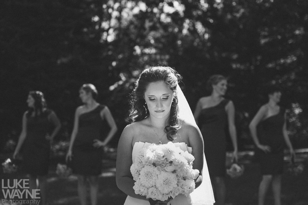 Bride in a white dress, holding a bouquet, with bridesmaids in dark dresses behind her. Black and white photo.