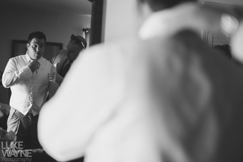 Man in a vest adjusting a tie in front of a mirror, with another person visible. Black and white photo.