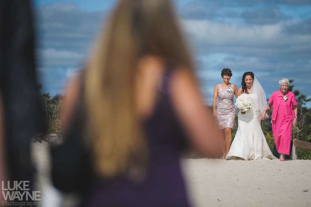 Bride walks down an aisle towards the camera, followed by bridesmaid and elderly woman in bright pink.