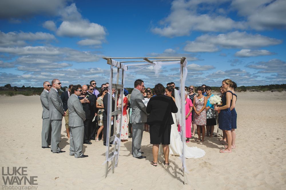 Wedding ceremony on a sunny beach with guests surrounding a couple under a decorated arch.
