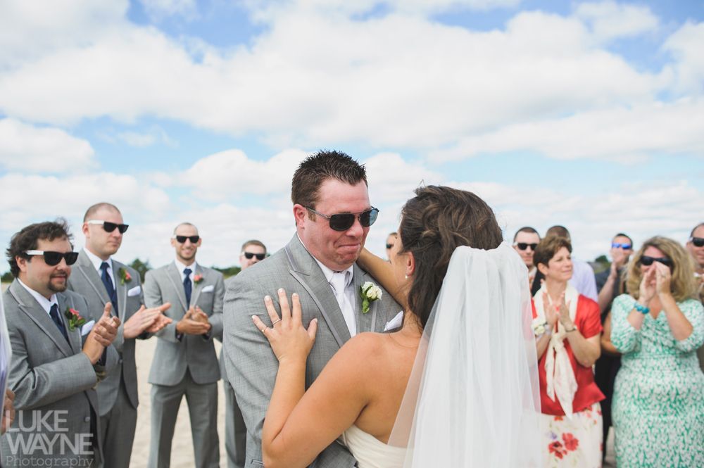 Wedding couple embracing on a beach, surrounded by guests in sunglasses and formal wear. Cloudy sky.