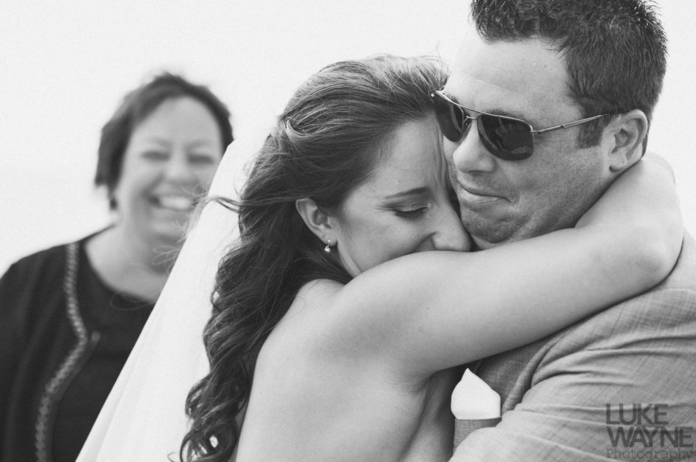 Bride hugging groom; woman smiles in the background. Black and white photo; wedding setting.
