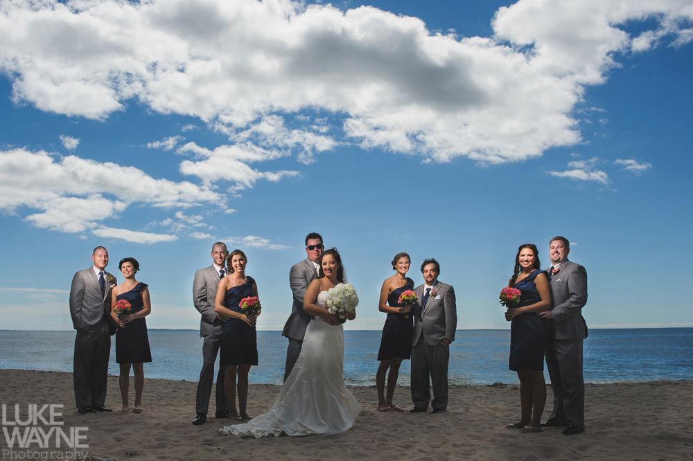Wedding party on a beach. Bride holding bouquet; bridesmaids wear navy, groomsmen gray. Blue sky with white clouds.
