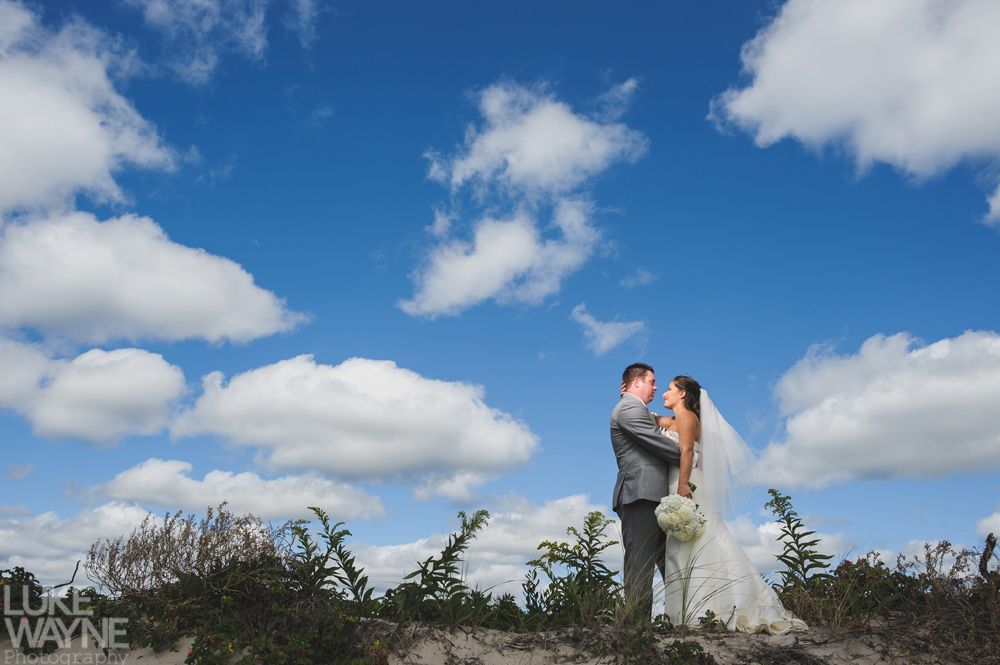 Bride and groom embrace beneath a bright blue sky filled with fluffy white clouds.