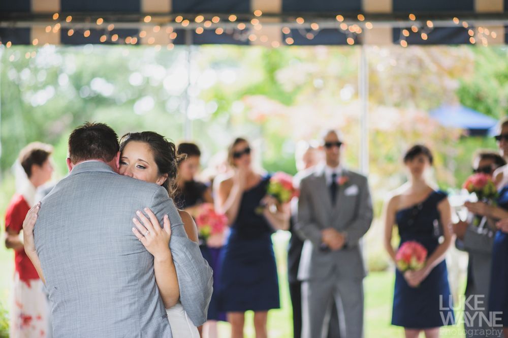 Bride and groom embrace during a wedding ceremony; bridesmaids and groomsmen watch, outdoor setting.