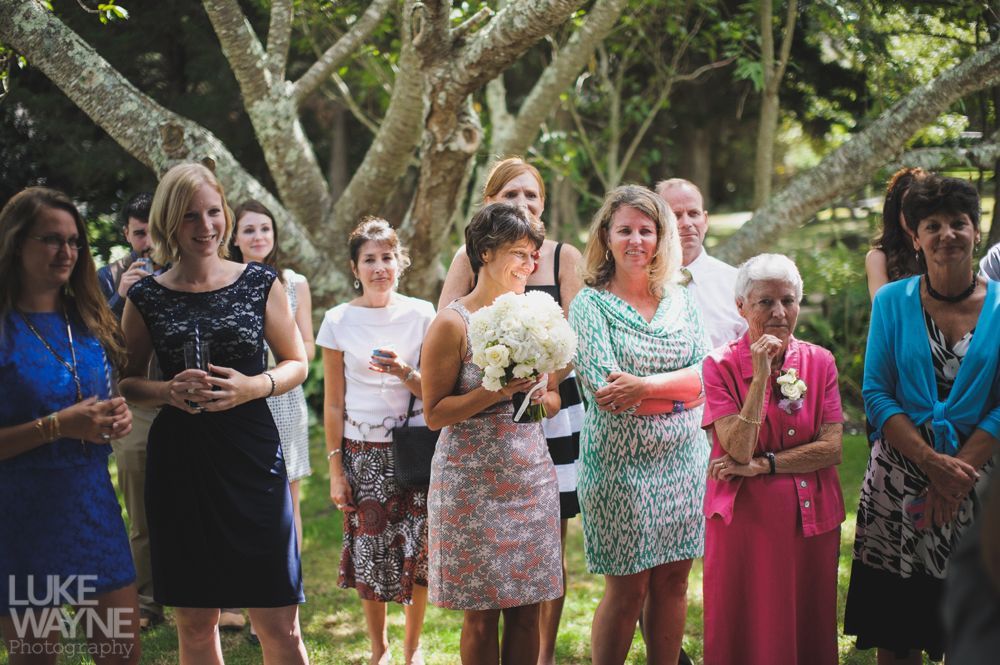 Guests watching a ceremony in a garden. Woman in pink dress holds bouquet. Trees provide shade.
