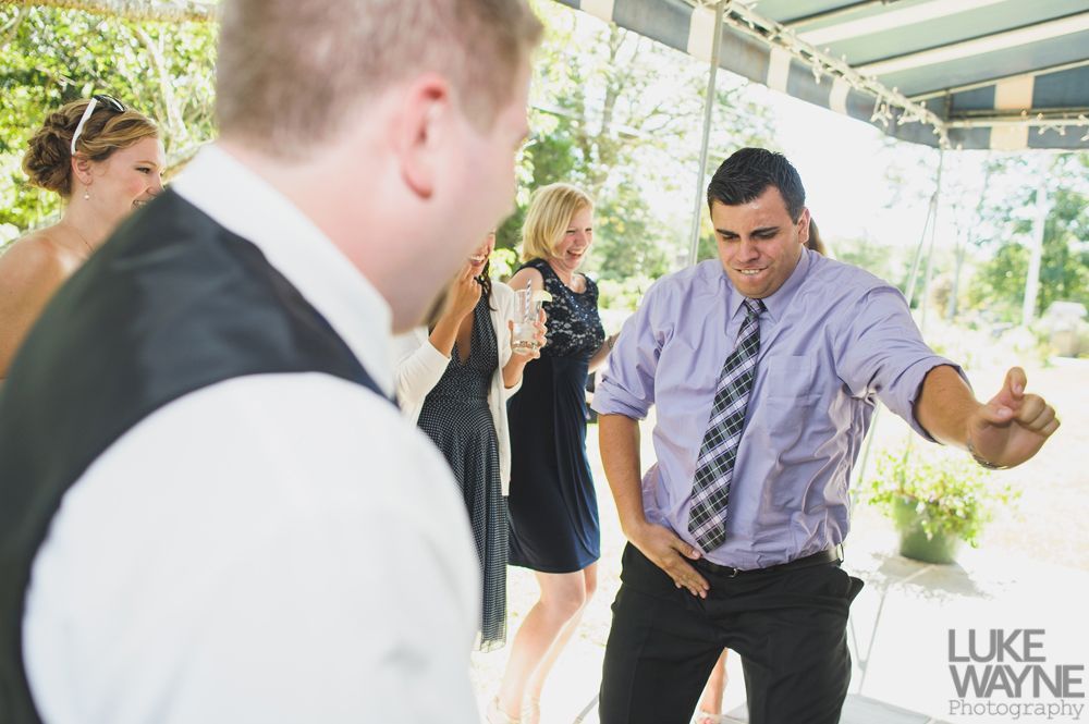 Man dances at an outdoor event, gesturing with arm extended, wearing a tie and dress shirt. Others watch and smile.