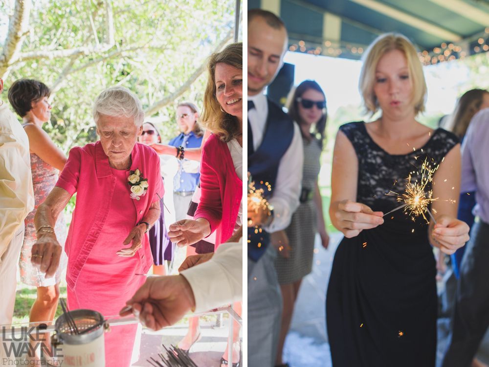 Wedding guests lighting sparklers. Elderly woman in pink dress; other guests watch. Woman in black dress with sparkler.
