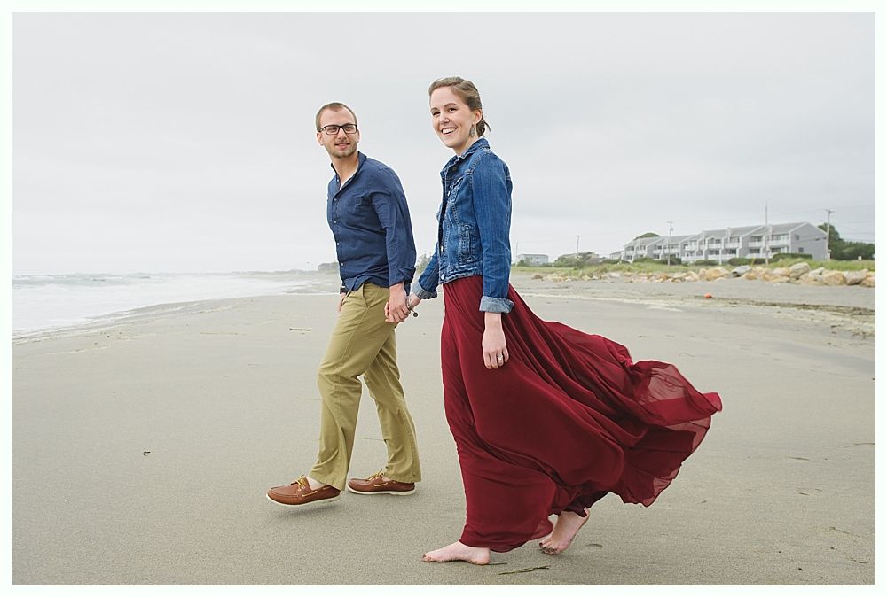 Couple walking on beach, holding hands, woman in red dress, man in blue shirt.