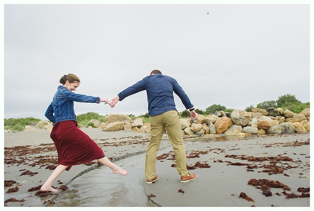 Couple holding hands, walking on a beach. Woman in red skirt, denim jacket, man in blue shirt, tan pants. Cloudy sky.