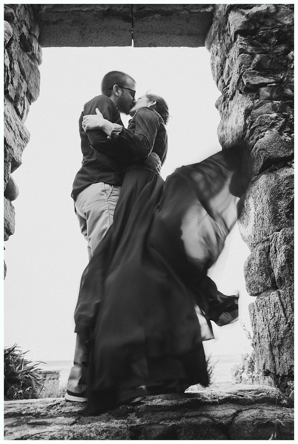 Couple embraces, kissing inside a stone window frame. Dress swirls. Overcast sky.