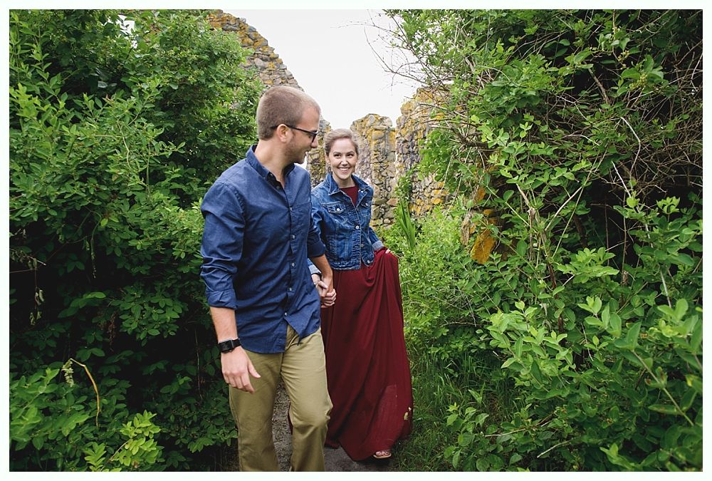 Couple holding hands, walking on a path surrounded by green bushes and stone walls. The woman wears a red skirt.