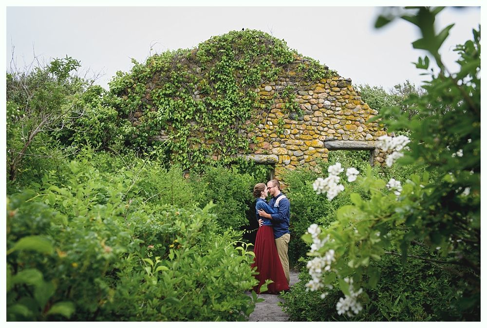 Couple embraces in front of a stone ruin overgrown with vines; lush green foliage surrounds them.