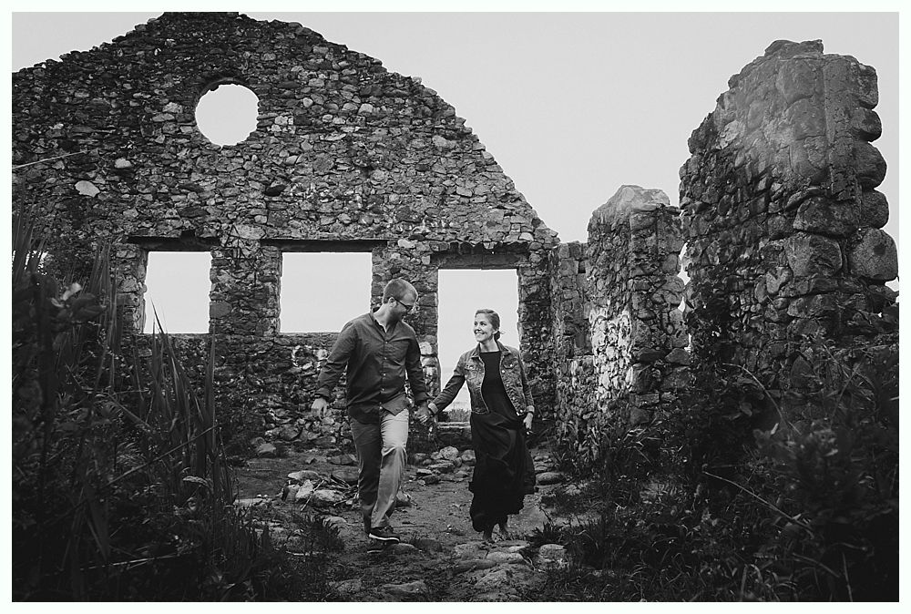 Couple holding hands, walking toward camera. Ruins of stone building with empty windows and a round opening in the wall behind them.