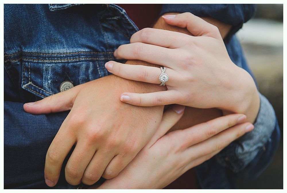 Hands clasped, one wearing a diamond engagement ring, against a denim jacket.