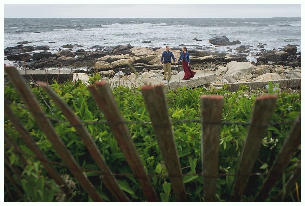 Couple holding hands walks by the rocky coast with ocean waves, seen through a wooden fence.
