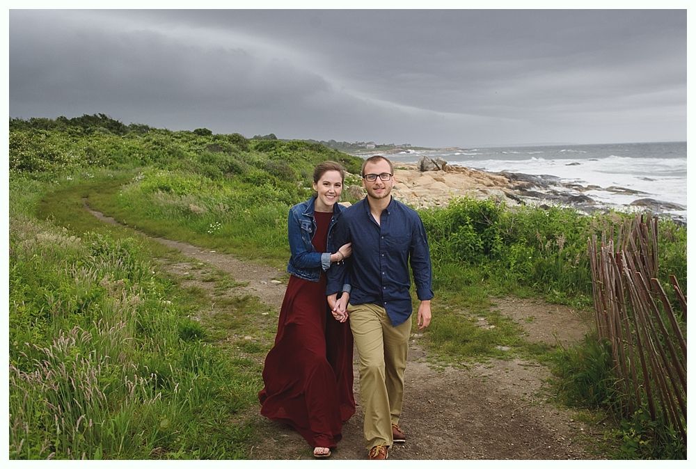 Couple walking hand-in-hand on a path near the ocean under a cloudy sky. Woman wears red dress and jean jacket. Man wears blue shirt and khakis.