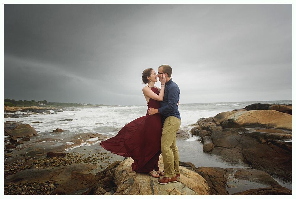 Couple embraces on rocks by the ocean; woman in burgundy dress, man in blue shirt. Stormy sky.