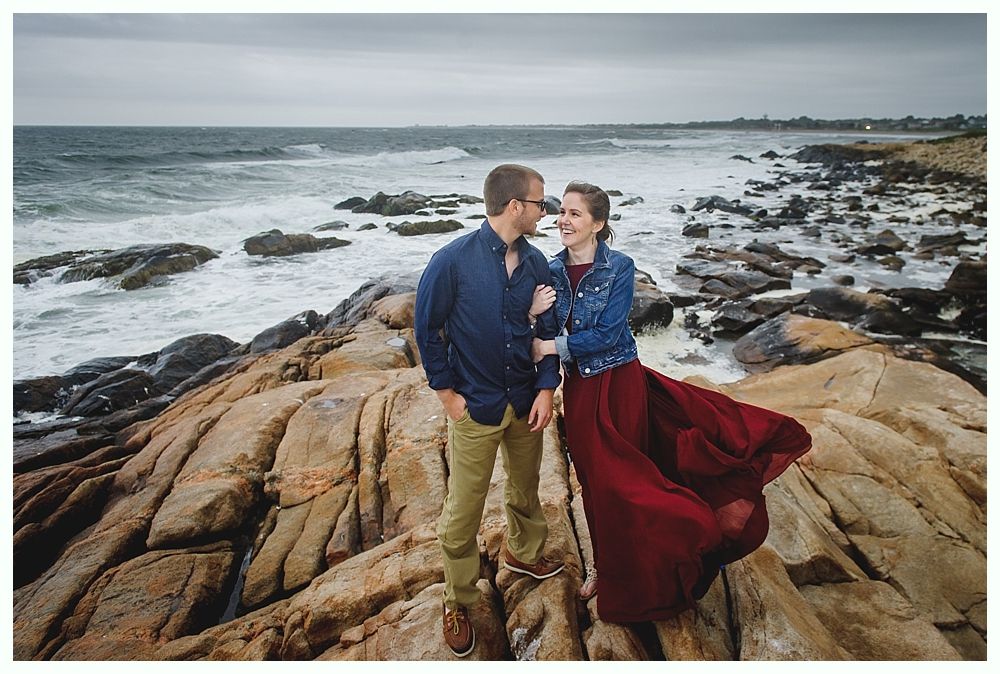 Couple embraces on rocky shore, woman in flowing red dress, man in blue shirt, ocean waves in background.