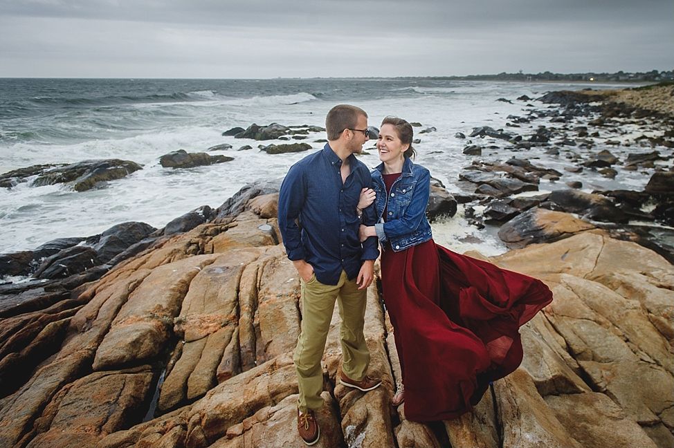 Couple standing close together on rocky shore by the ocean. She wears red dress and denim jacket. He wears blue shirt and khaki pants.