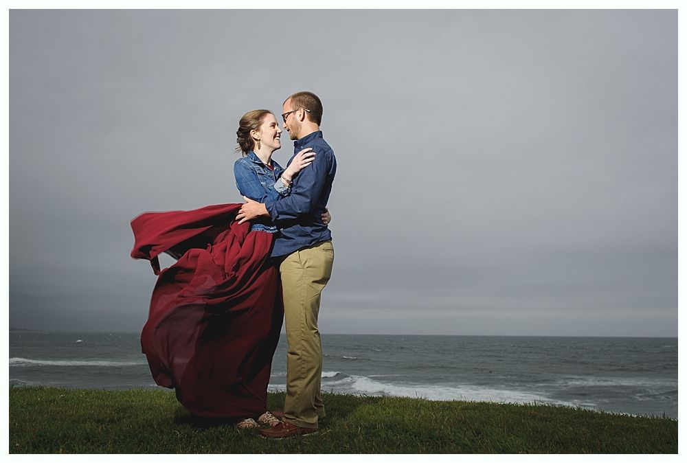 Couple embraces by the ocean; woman's red skirt billows in wind, stormy sky.